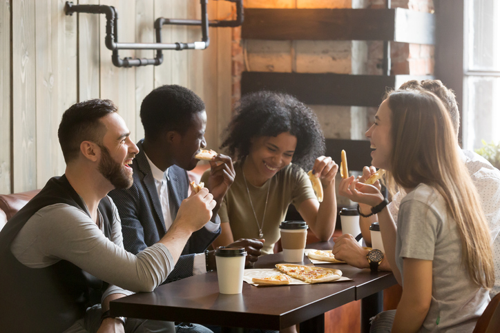 Multiracial happy young people eating pizza in pizzeria, black and white cheerful mates laughing enjoying meal having fun sitting together at restaurant table